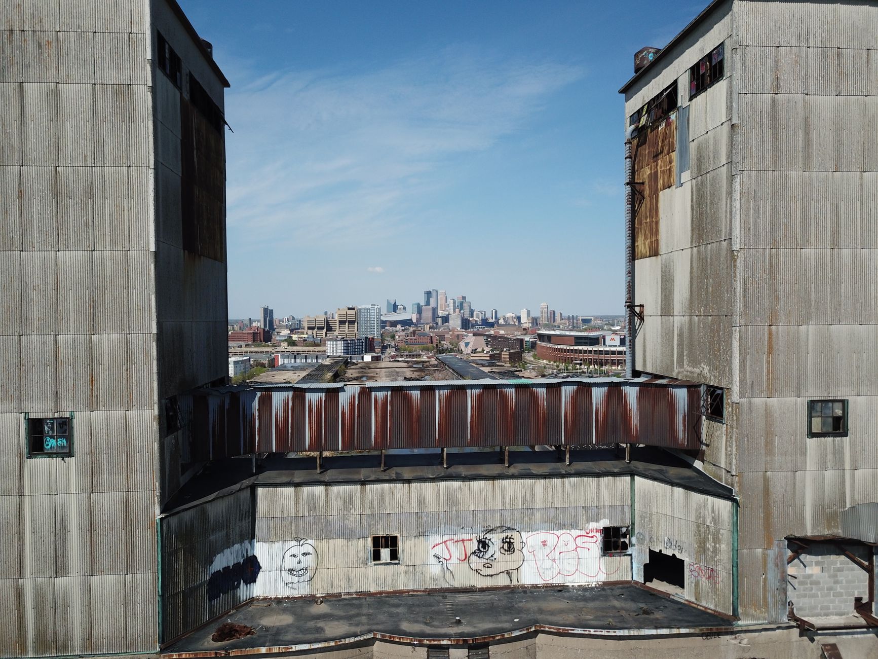 A rusted grain elevator tower rises against a gray Minneapolis sky with a chain-link fence in the foreground.